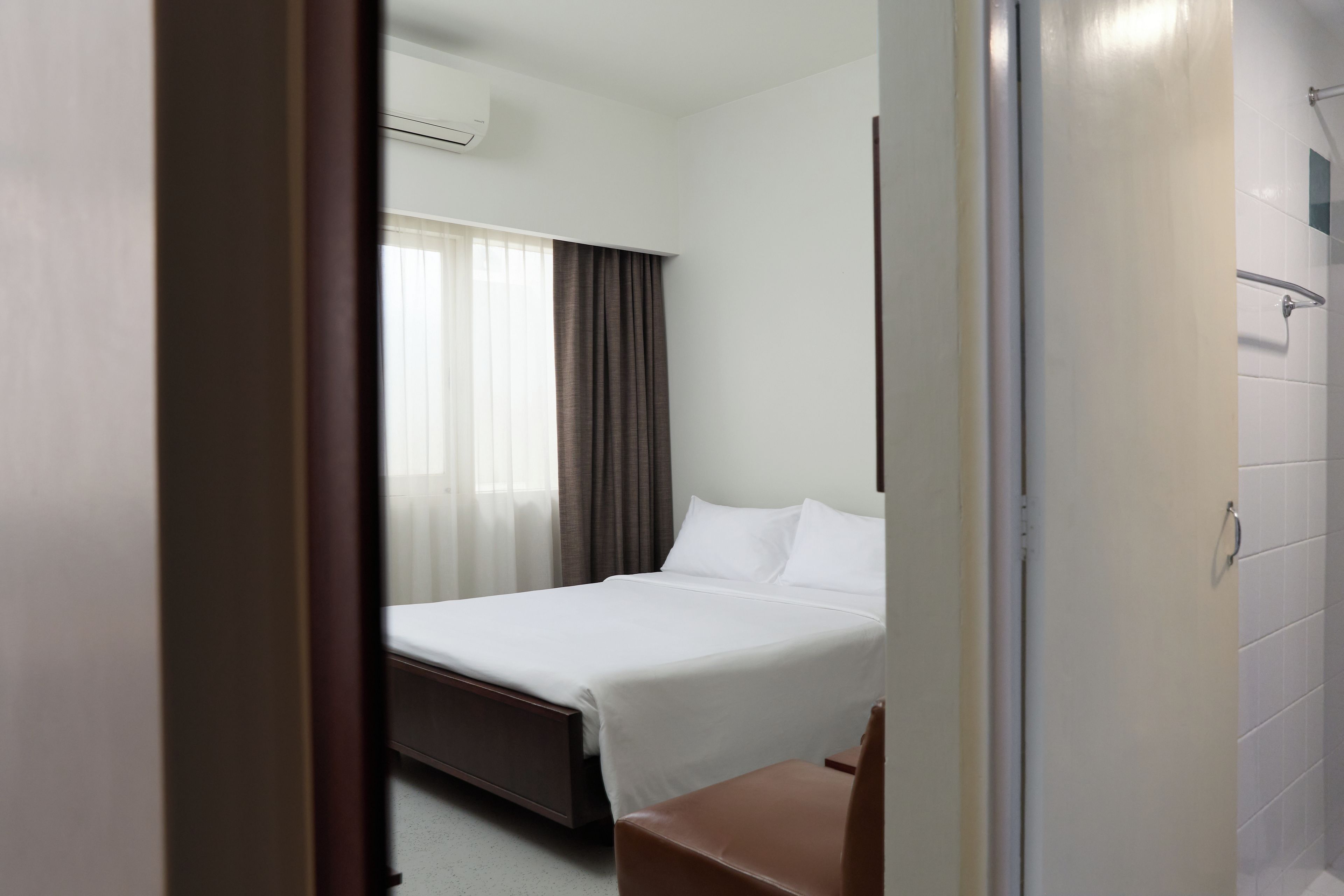 A hotel room with a bed, a chair, a window with curtains, and a shower area.
