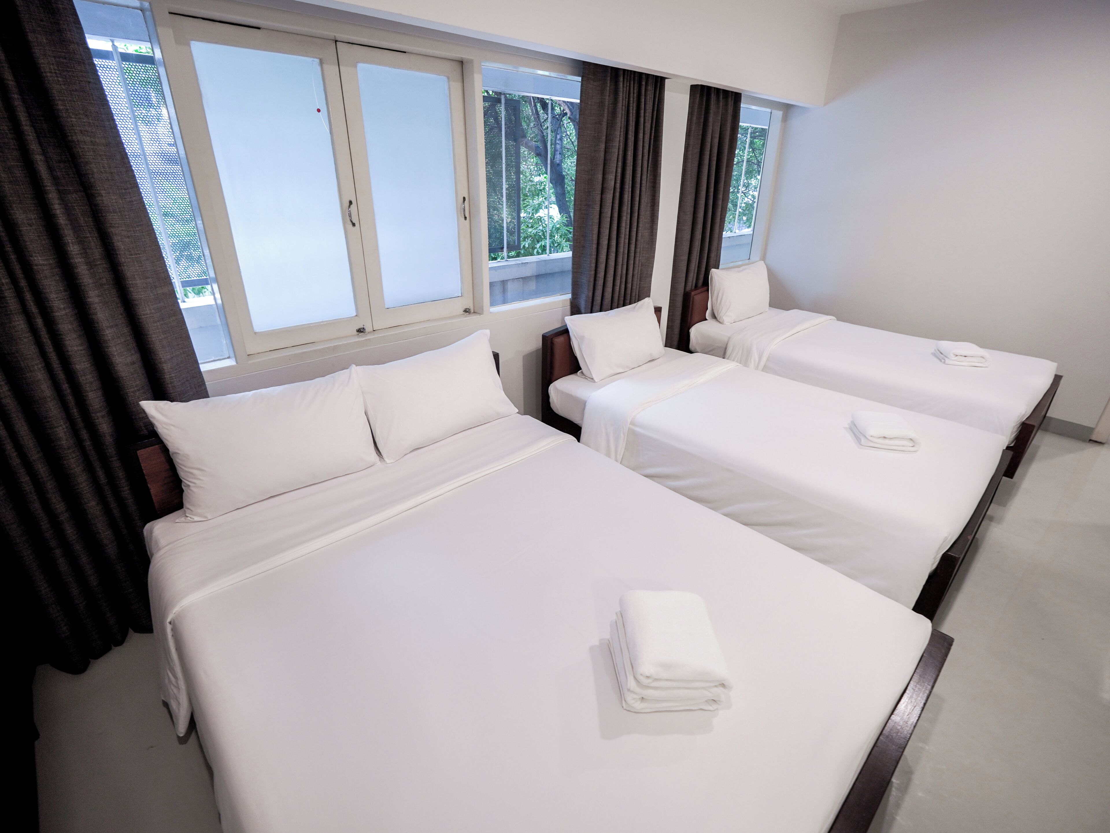 Quarto de hotel com três camas de solteiro, uma janela grande com cortinas e vista para o verde lá fora.