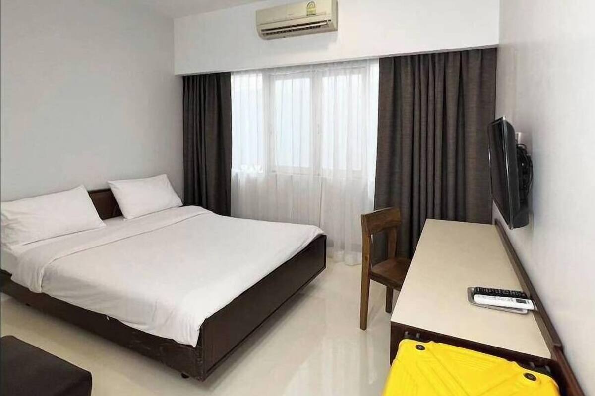A hotel room with a bed, a desk, a chair, a TV, and an air conditioning unit.