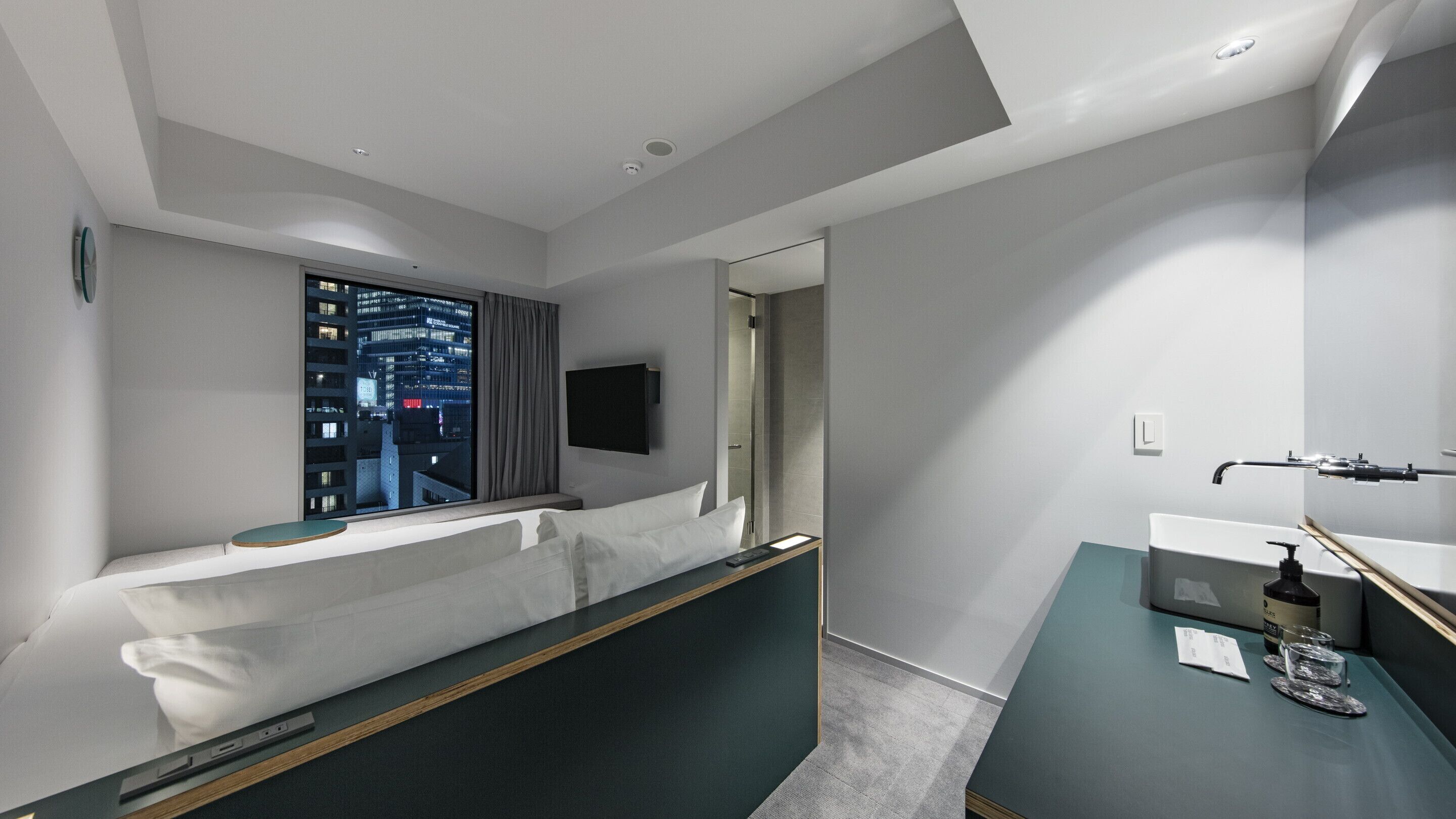 A modern hotel room with a large window offering a city view, a bed with a headboard, a bedside table with a bottle and glasses, and a small desk area.