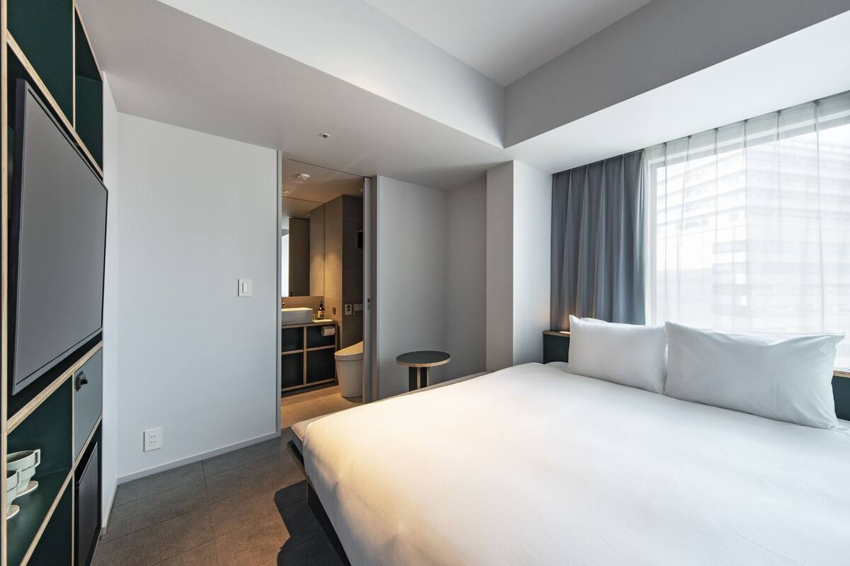A hotel room with a large bed, a desk, a chair, a wardrobe, and a bathroom visible through an open door.