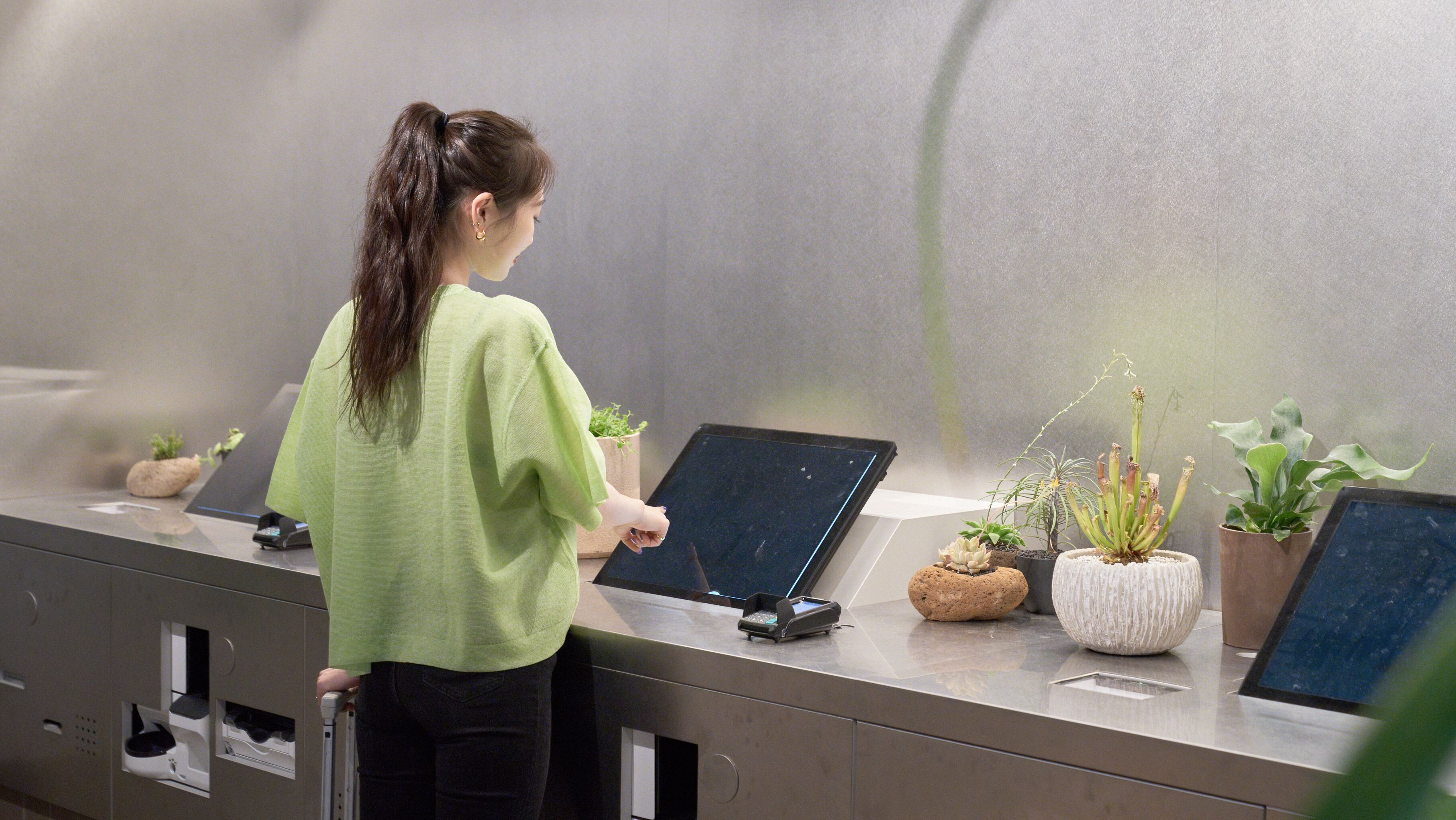 A woman at a self-service kiosk with multiple screens and potted plants.