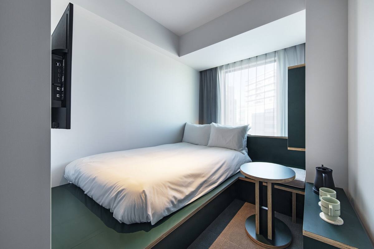 A compact hotel room with a bed, a small round table with a kettle and cups, a window with curtains, and a wall-mounted TV.