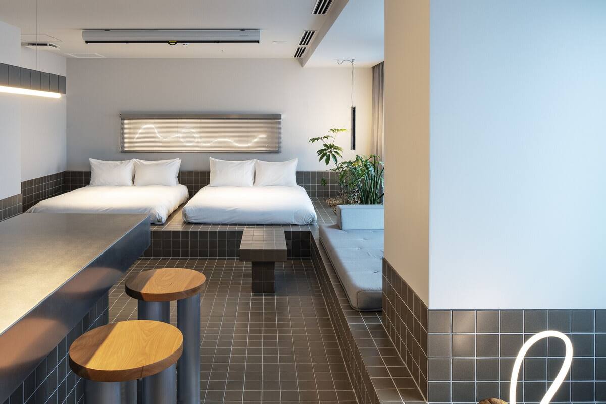 A modern hotel room with two beds, a small seating area, and a large window with blinds.