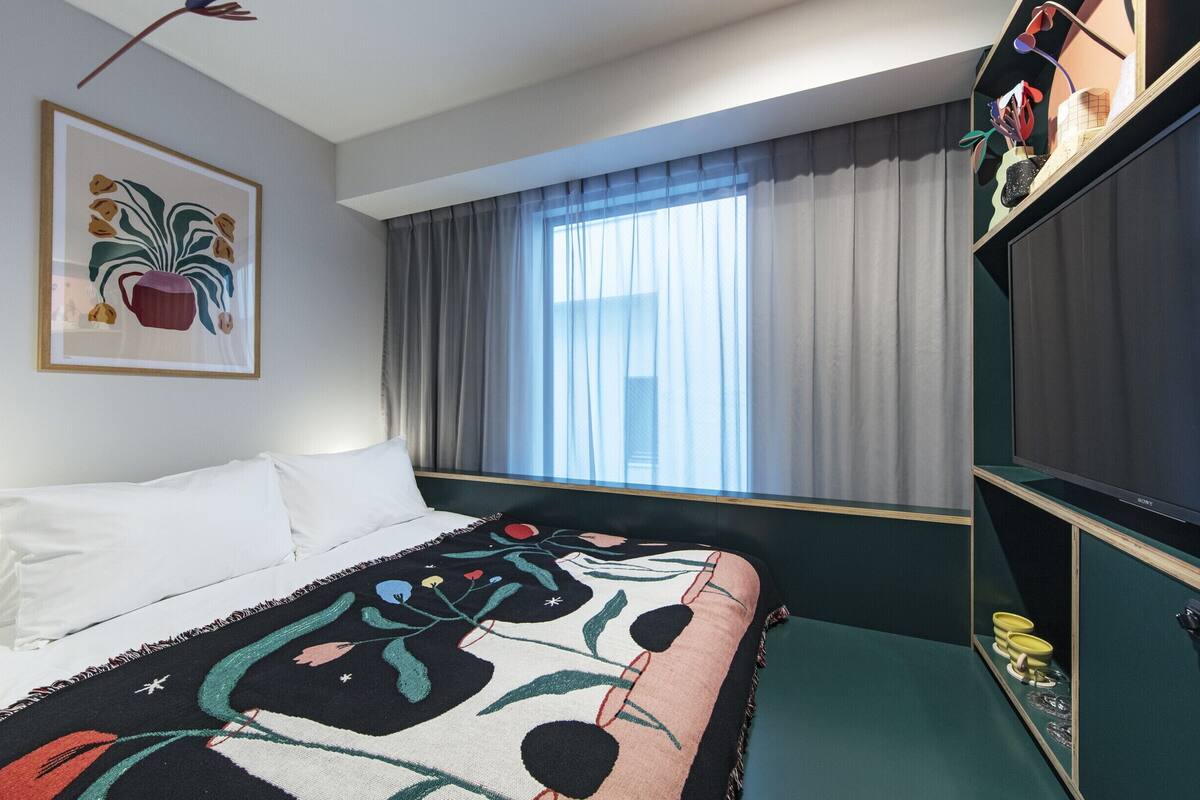 A hotel room with a bed, a large flat-screen TV, a window with curtains, and a framed artwork on the wall.