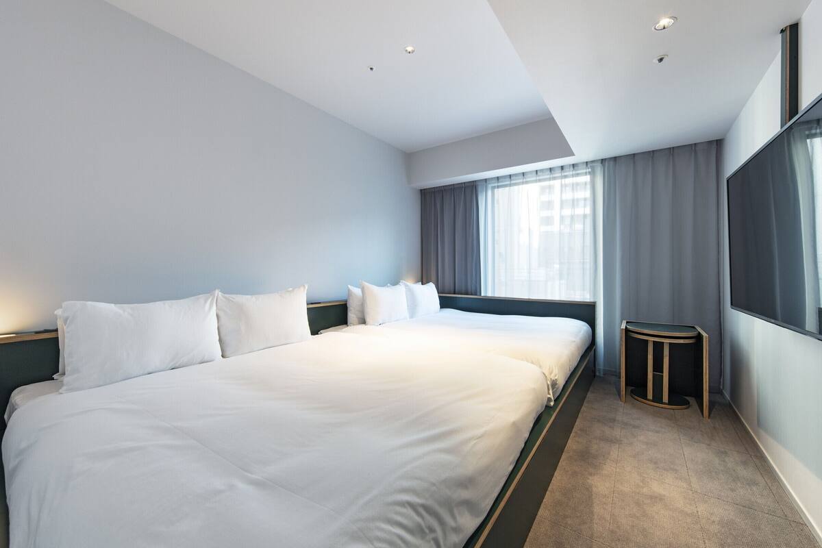 A modern hotel room with a large bed, a flat-screen TV, a small side table, and a window with curtains.