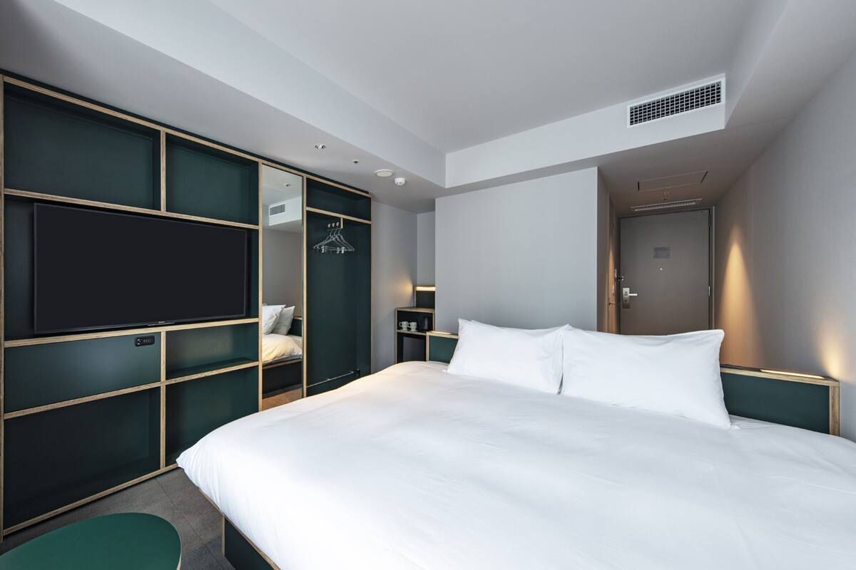 A hotel room with a large bed, a flat-screen TV mounted on a dark green wall, a wardrobe with a mirror, and a door with a handle.