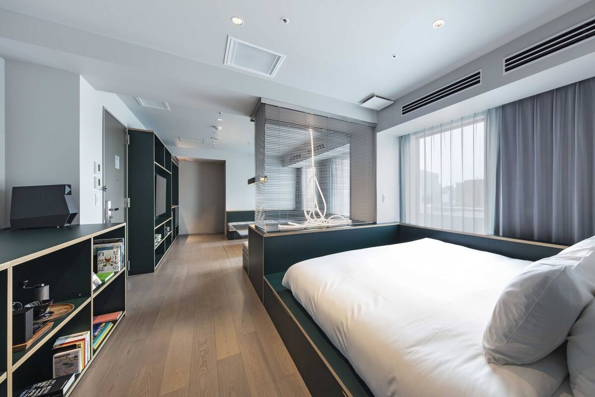 A modern hotel room with a large bed, a seating area, a bookshelf, and a television.