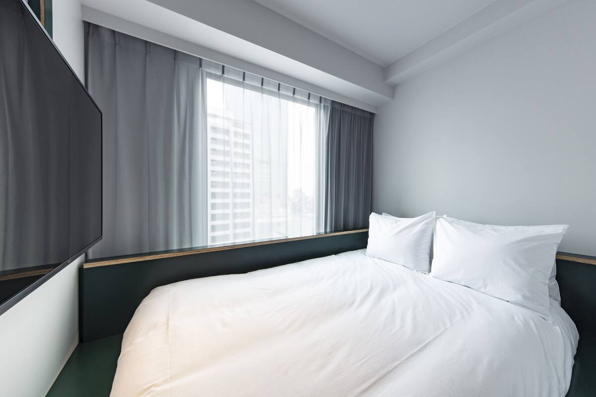 A hotel room with a bed, a window with curtains, and a view of buildings outside.