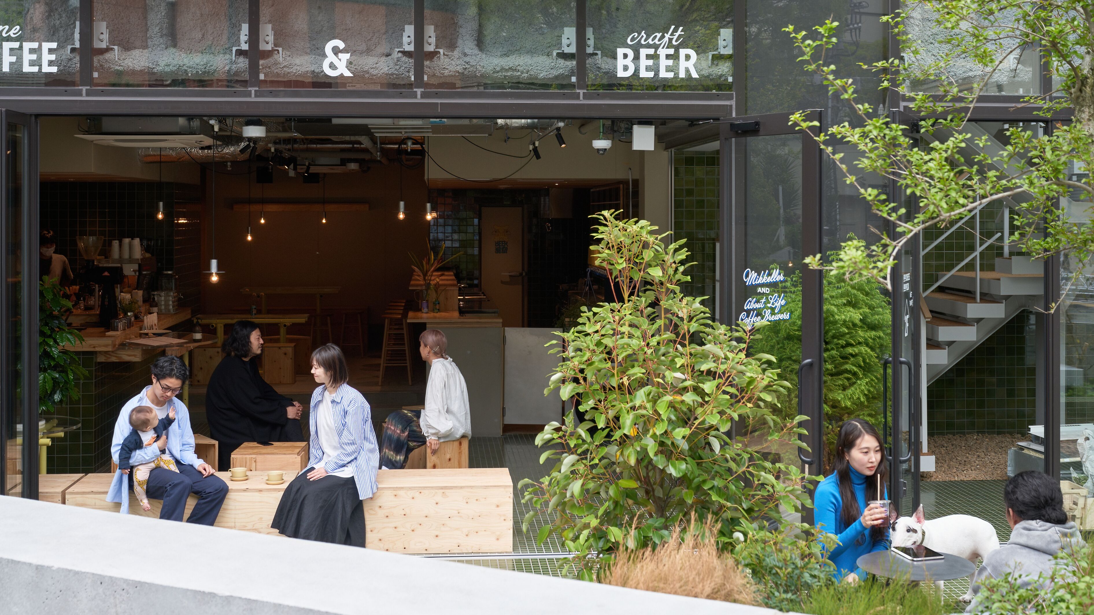 Et udendørs siddeområde foran en café med moderne design, inklusive en trappe og et lille træ.