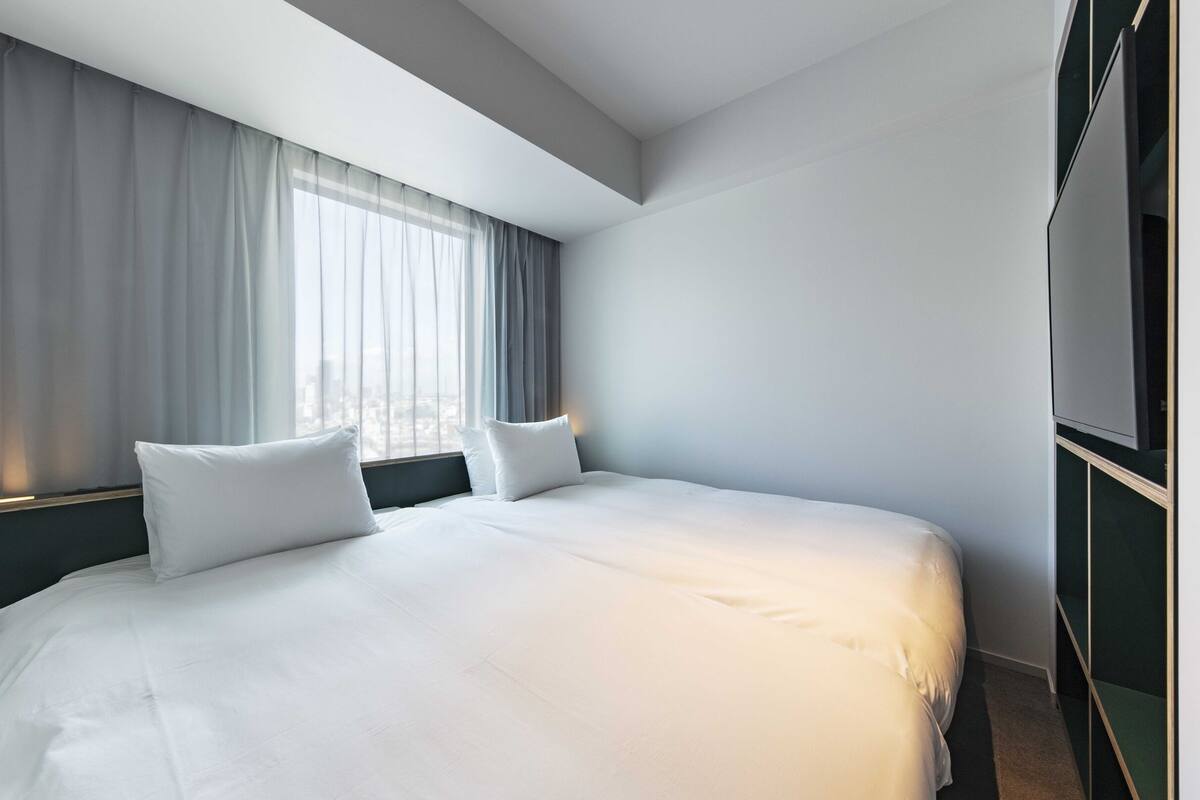 A modern hotel room with a large bed, two pillows, a dark headboard, a large window with sheer curtains, and a flat-screen TV mounted on the wall.