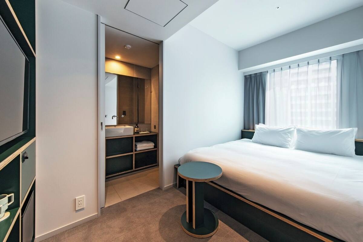 A modern hotel room with a large bed, a small round table, a built-in wardrobe, a bathroom with a mirror and sink, and a large window with curtains.