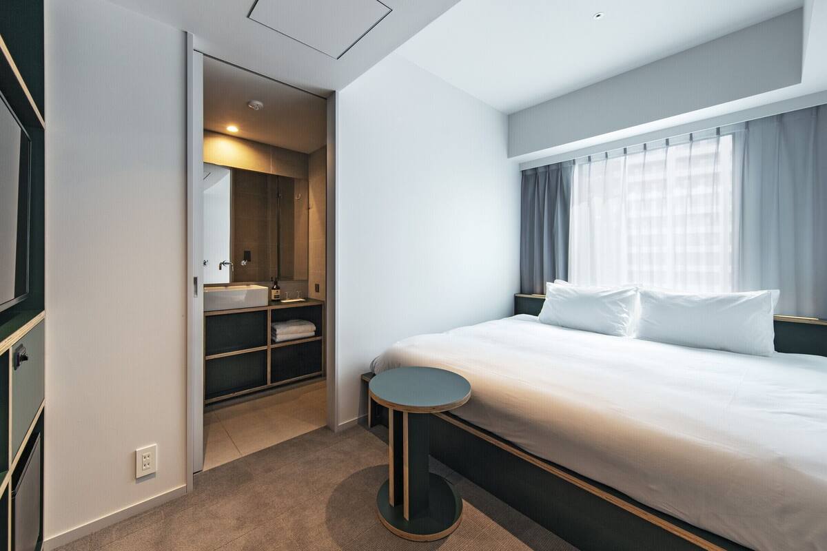 A modern hotel room with a large bed, a walk-in closet, a bathroom with a mirror, and a window with curtains.