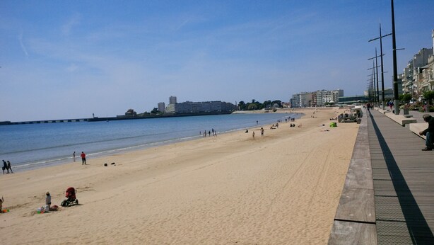 A wide sandy beach with a promenade on the right, a calm sea, and buildings in the background. - For family fun at the beach, at the center of everything that matters (Les Sables-d'Olonne)