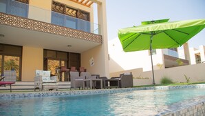 A swimming pool with tiled edges, outdoor furniture, and a green umbrella. - Villa-Bay (Muscat)