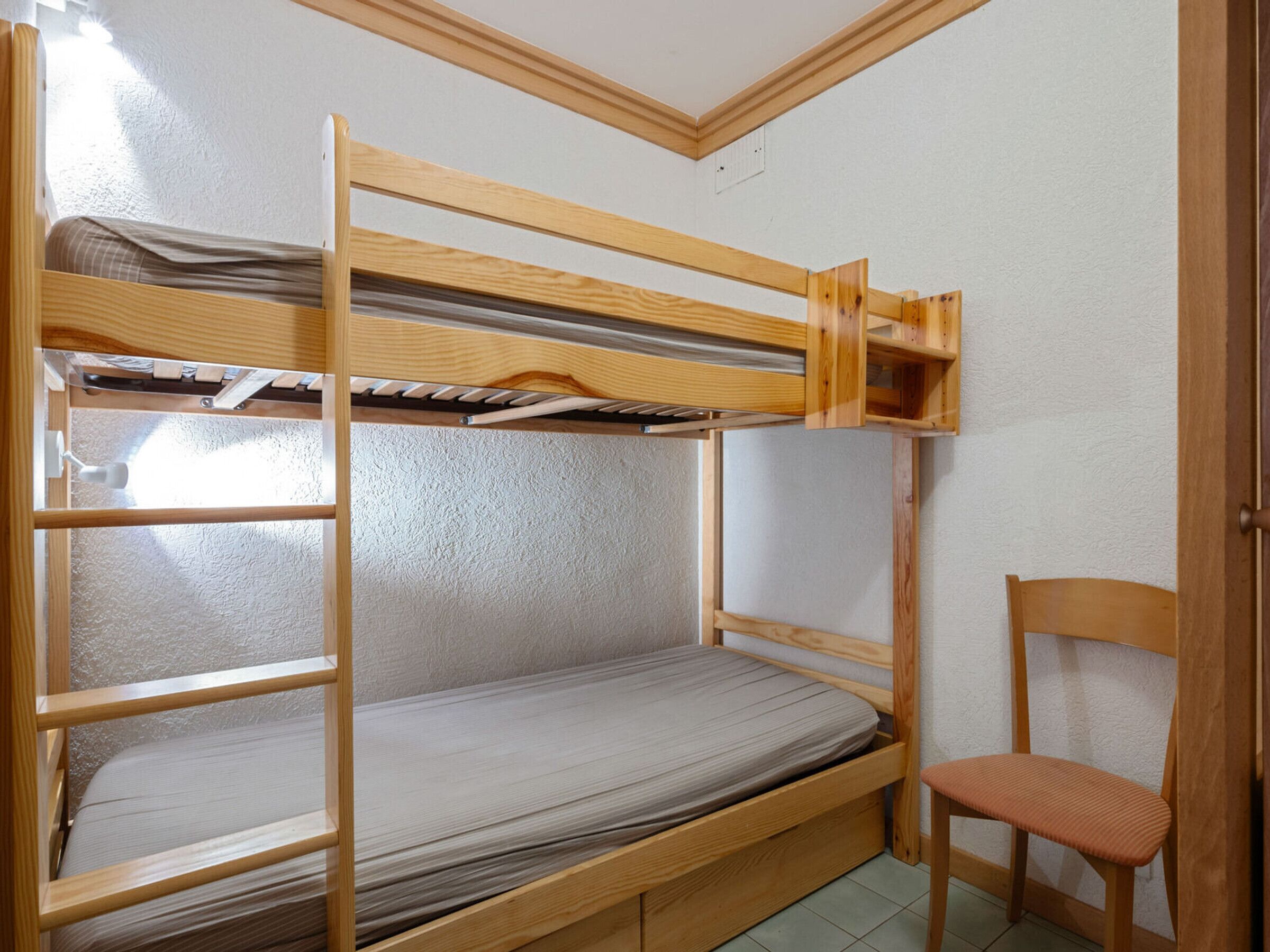Una cama litera con estructura de madera, una cama individual abajo y una silla a la derecha.