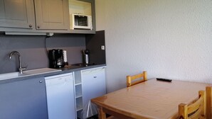 Kompakti keittiö, jossa on harmaat kaapit, mikroaaltouuni, kahvinkeitin ja astianpesukone. Keittiössä on puinen pöytä, jonka päällä on tuoli ja kaukosäädin.