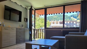 A modern living room with a sofa, a coffee table, and a TV mounted on the wall. There is a balcony with a view of a forested area and buildings. - Renovated studio 4/5 ppl, south balcony, private parking, Pra Loup (Pra-Loup)