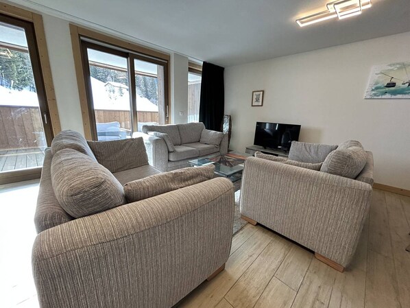 Una sala de estar con dos sofás, una mesa de centro, un televisor y ventanas grandes.