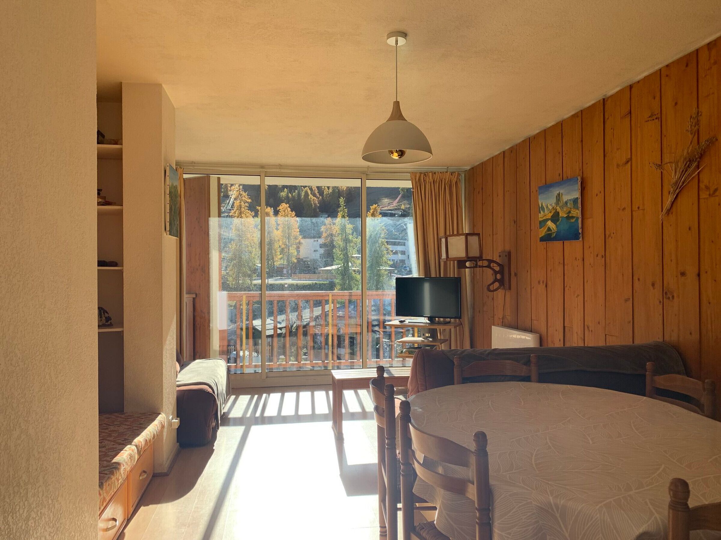 A room with a balcony, a television, a dining table, and a painting on the wall.