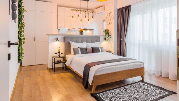A modern bedroom with a large bed, a bedside table with a lamp, and a view of a kitchen area. - Studio 54 by MRG Apartments (Bucharest)
