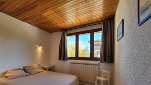 A bedroom with a wooden ceiling, a bed, a radiator, a window with curtains, and a chair.