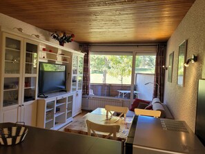 Espaço compacto com televisão, armários brancos, mesa de jantar e vista para o exterior.