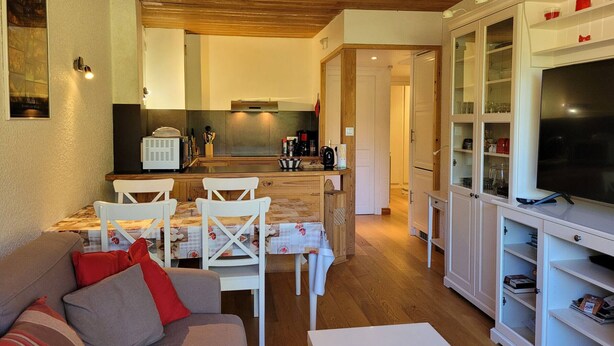 A cozy living room with a sofa, a dining area with chairs, a kitchen with a stove, and a TV in a cabinet.