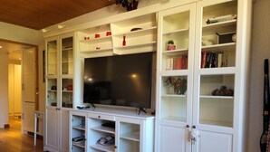 A living room with a large flat-screen TV on a white entertainment unit, surrounded by open shelving with books and decorative items.