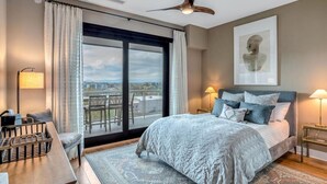 A bedroom with a large bed, a desk, a chair, a balcony with a view, and a ceiling fan.