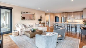 A modern living room with a sofa, armchairs, and a coffee table. There is a kitchen area with bar stools and pendant lighting.