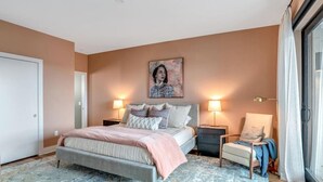 A bedroom with a bed, two bedside tables, a chair, a large painting on the wall, and a window with curtains.