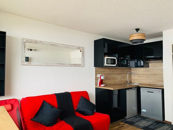 A compact kitchen with a microwave, sink, and dishwasher. A red sofa with black cushions is positioned in front of the kitchen.