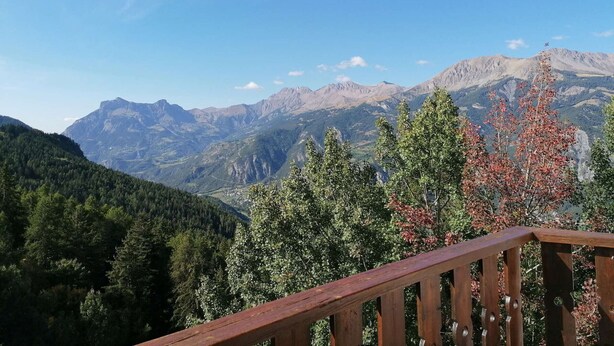 Vista de la montaña desde una terraza de madera con barandilla, que domina un valle con bosques densos y montañas lejanas.