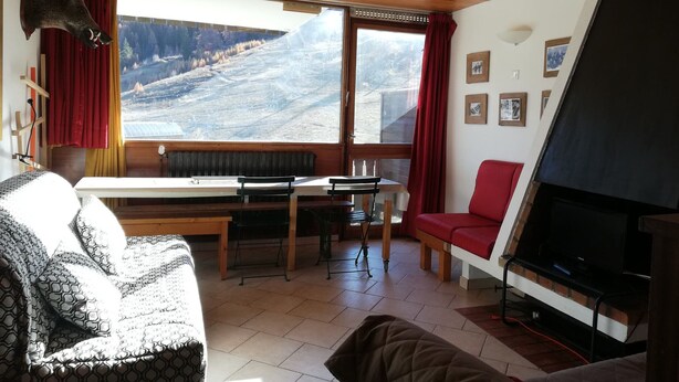Een gezellige kamer met een groot raam, een eettafel, een rode stoel, een bank en een open haard.