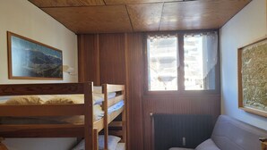 Een kleine kamer met een stapelbed, een slaapbank, een radiator en een raam met gordijnen.
