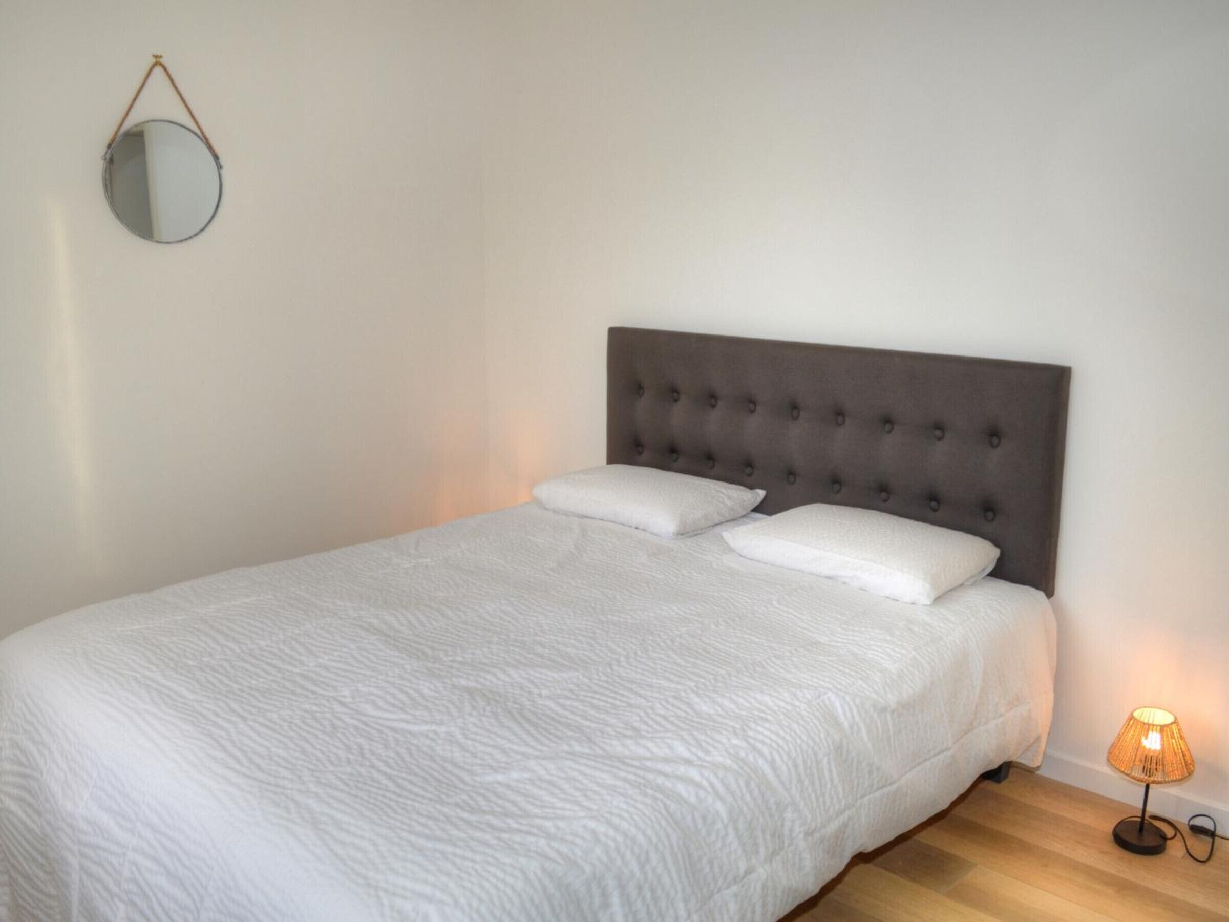 A neatly made bed with a tufted headboard, a round mirror on the wall, and a small table with a lamp beside it.