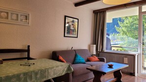 Una habitación acogedora con sofá, una mesa y un banco. Hay una ventana con cortinas y vistas a árboles fuera.