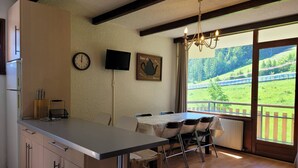 Une cuisine dotée d’un grand îlot central, un coin repas avec une table et des chaises, une horloge, un téléviseur et une vue sur un paysage verdoyant par la fenêtre.