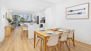 Uma sala de jantar moderna com uma mesa de madeira preparada para seis pessoas, cadeiras brancas e vista para a cozinha e a sala de estar.