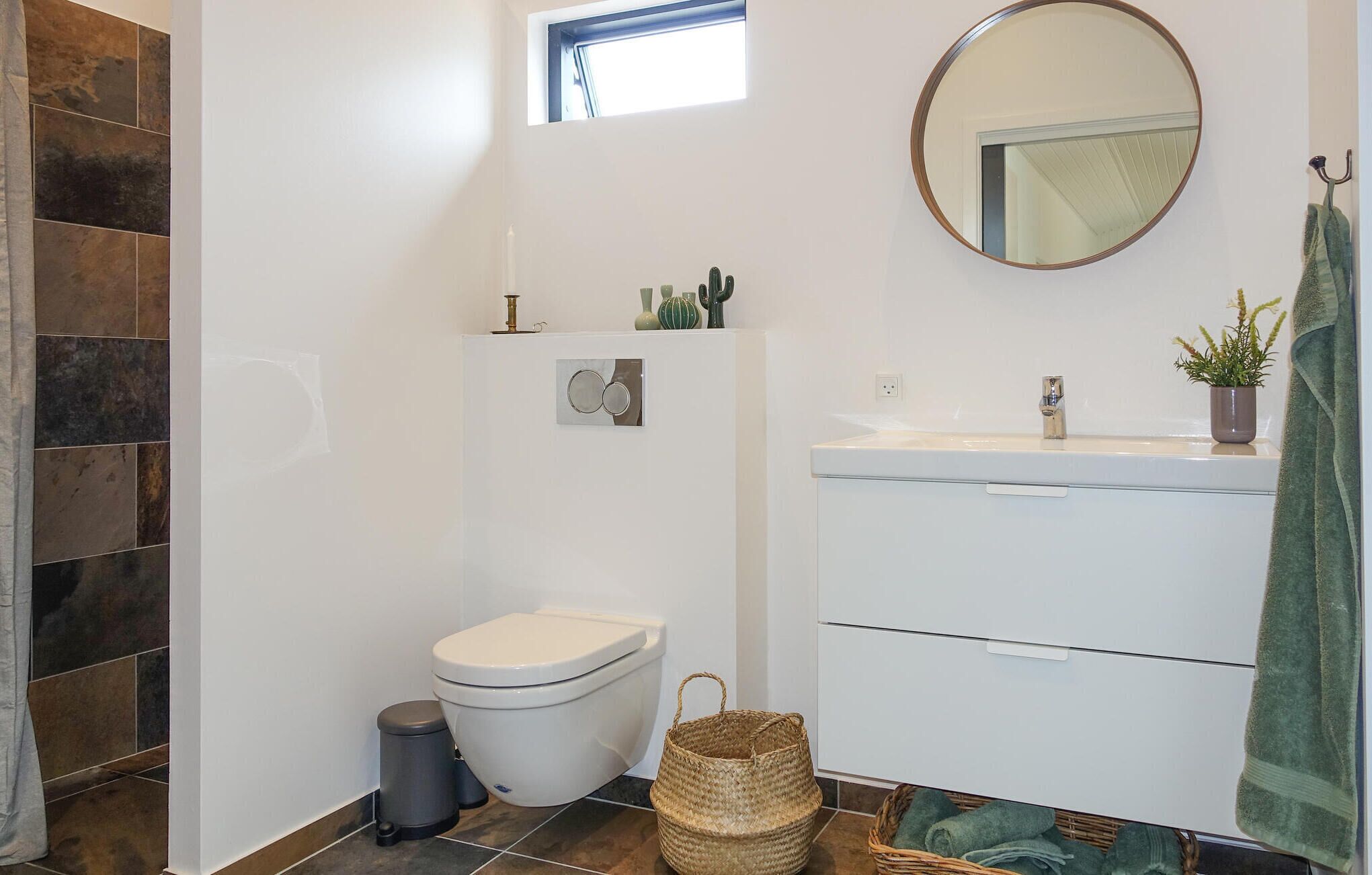 Ein modernes Badezimmer mit Toilette, Waschbecken und Spiegel. Über der Toilette befindet sich ein kleines Fenster und auf dem Boden ein Wäschekorb mit Handtüchern.
