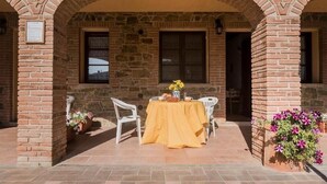 Une terrasse extérieure avec une table recouverte d’une nappe jaune, entourée de chaises blanches, et un mur de briques surmonté d’une arche en pierre.