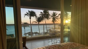 Une chambre d’hôtel dotée d’un grand lit, d’un balcon offrant une vue sur l’océan et les palmiers, ainsi que d’un coin repas avec des sièges en plein air.