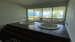 Une salle de bain moderne avec deux jacuzzis sur une plateforme en bois, une grande fenêtre donnant sur la mer et une inscription au sol indiquant « COSTA ESPAÑA ».