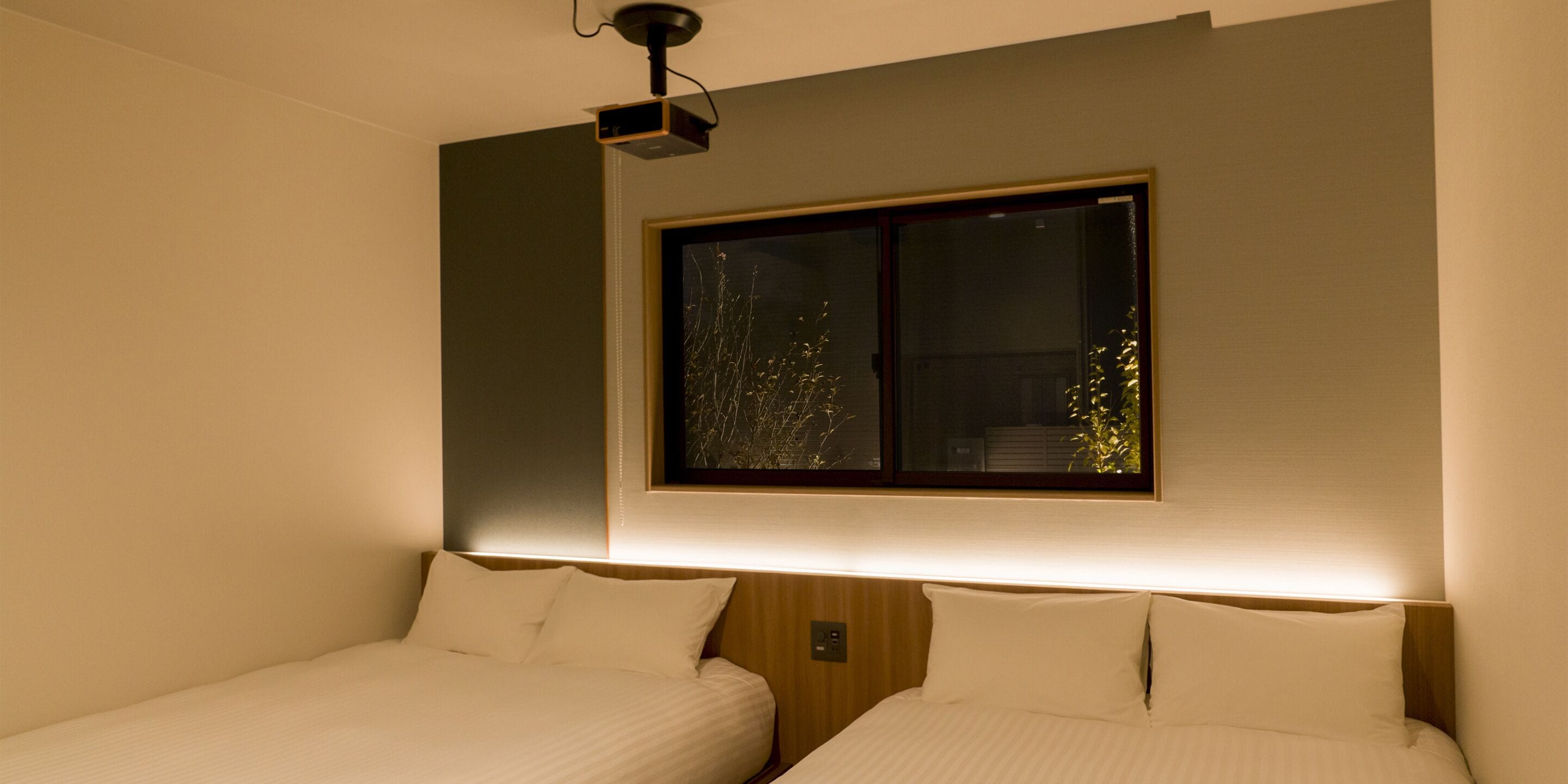 A hotel room with two beds, a ceiling projector, and a window with a view of trees.