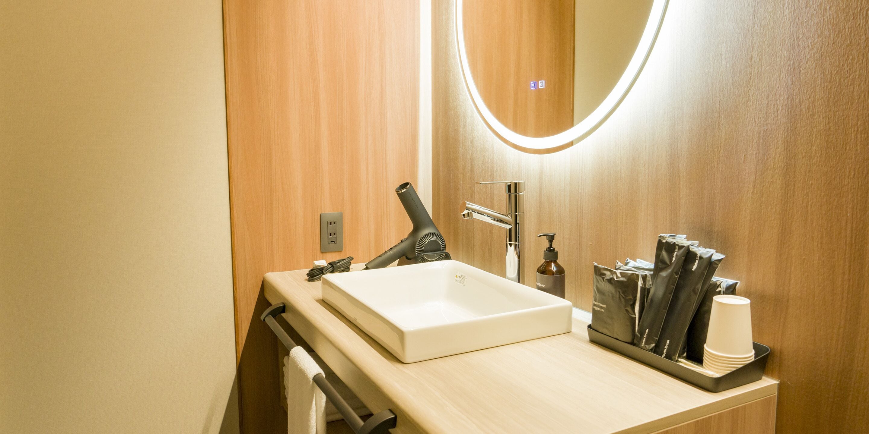 Ein modernes Badezimmer mit einem Holzwaschtisch, einem weißen Waschbecken, einem Spiegel mit Beleuchtung, einem Fön und einem Handtuchhalter.