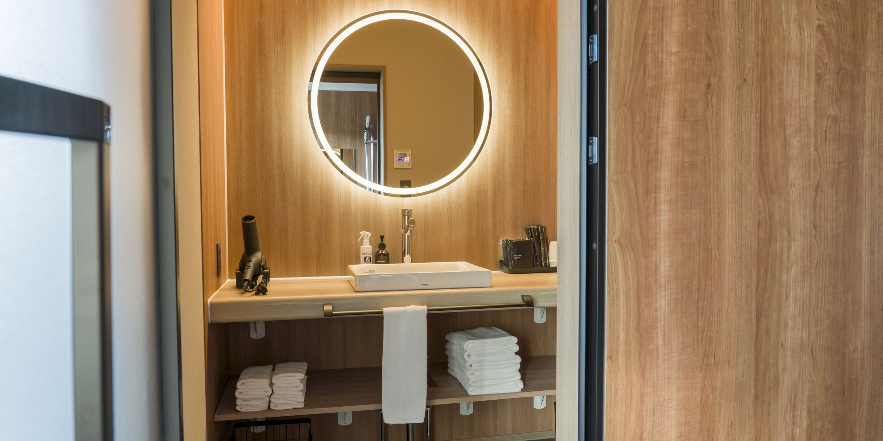 Une salle de bain moderne avec un meuble-lavabo en bois, un miroir rond doté d’un éclairage et des serviettes soigneusement pliées, rangées sur des étagères.