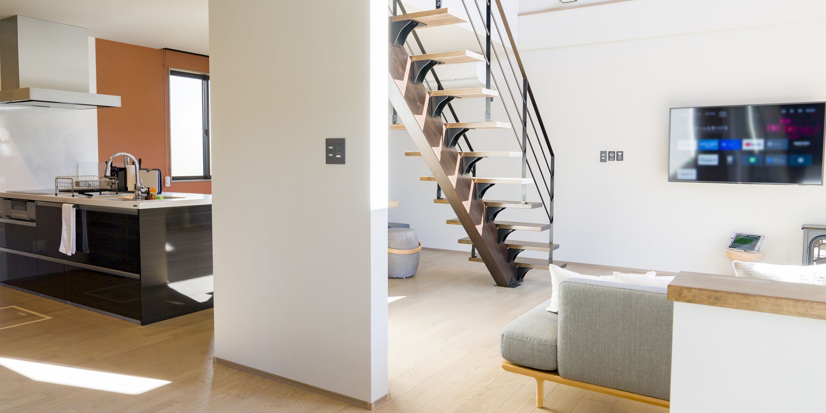 A modern kitchen with a built-in oven and a dining area with a grey sofa and a wooden staircase.
