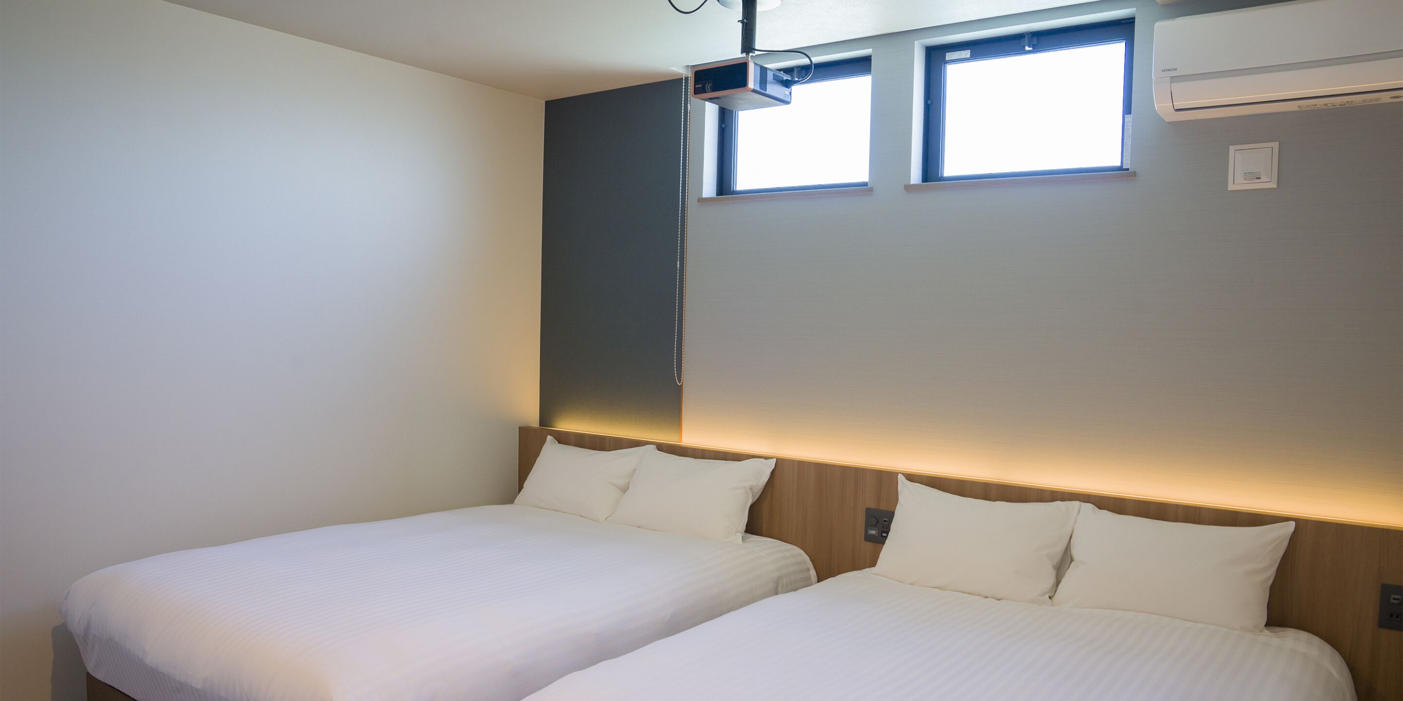 A hotel room with two beds, a ceiling-mounted projector, and an air conditioning unit.
