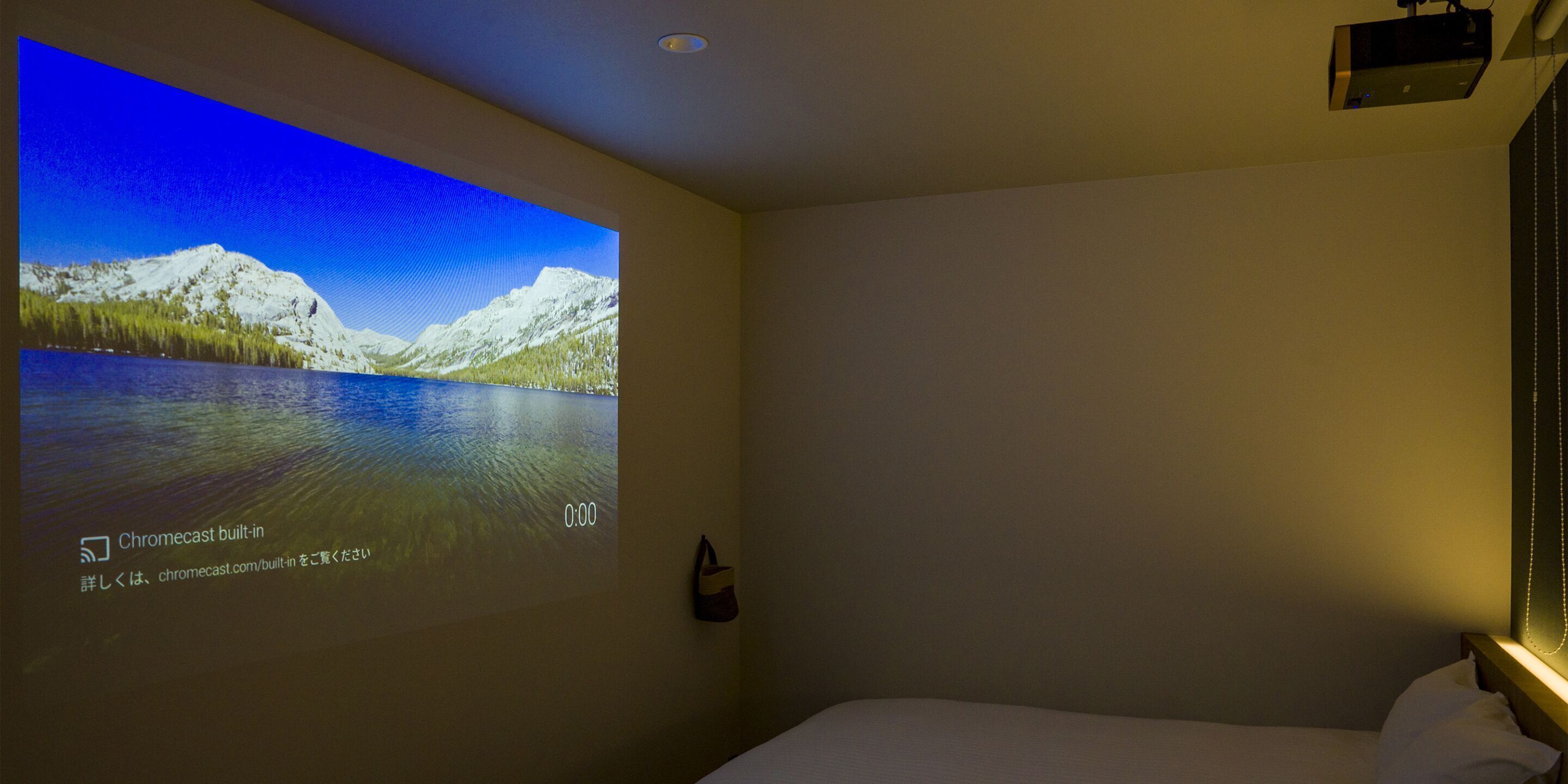 A hotel room with a bed, a wall-mounted projector displaying a scenic image, and a small nightstand with a lamp.